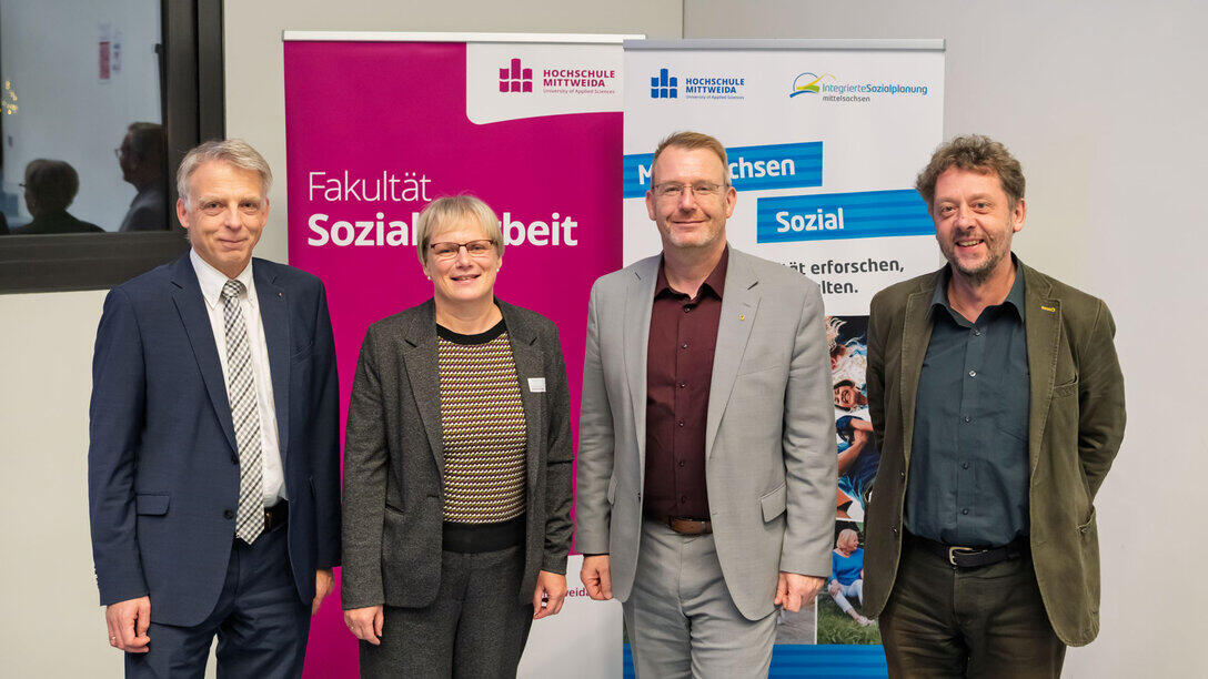 A group photo showing three men and one woman at the far end of the lecture theatre in front of roll-up banners reading ‘Faculty of Social Sciences’ and ‘Mittelsachsen Sozial’."