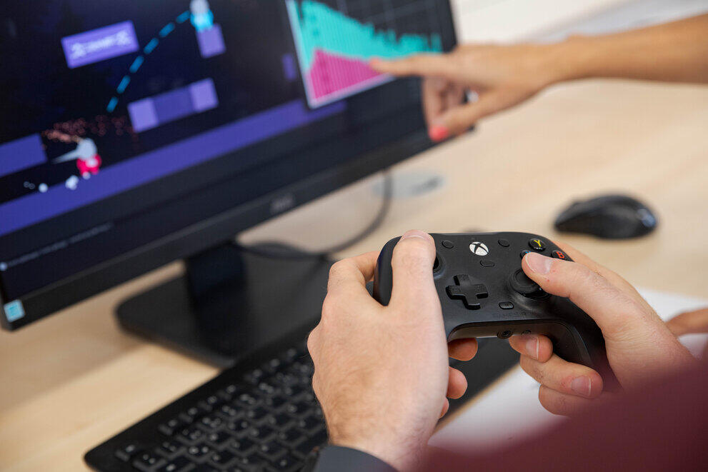 A student is holding an Xbox controller in front of a monitor on which a video game editor is running.