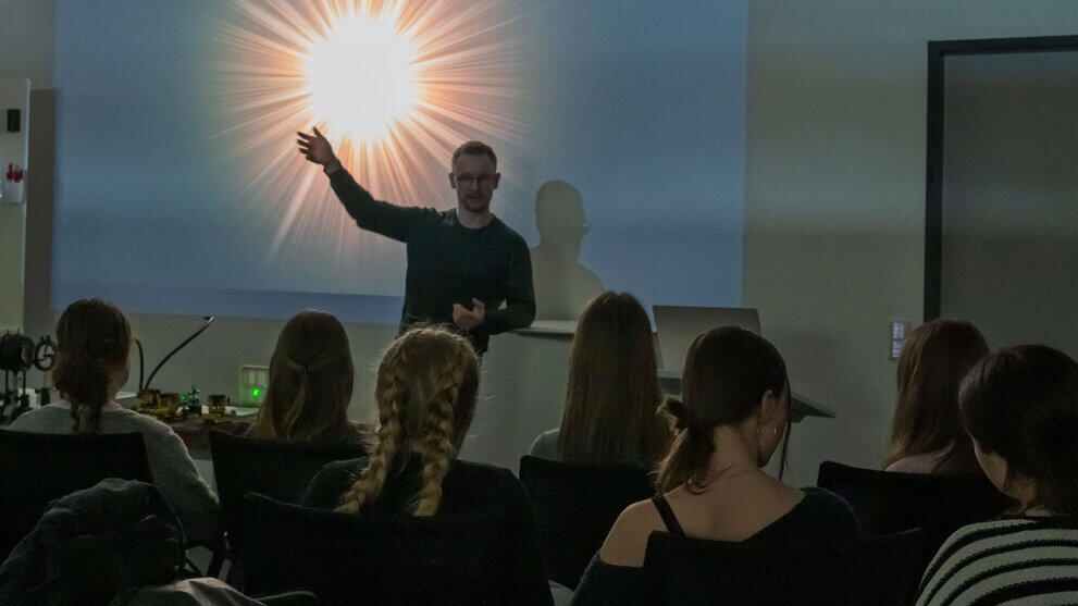 Blick in einen abgedunkelten Seminarraum über die Köpfe von Schüler:innen hainweg auf einen Dozenten, der vor einer hell leuchtenden Projektion an der Wand steht.