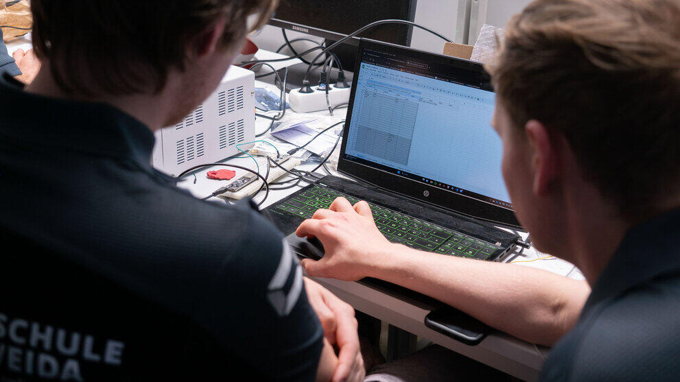 Aufgeklappter Laptop, vor dem zwei männliche Personen sitzen. Auf dem Display ist eine Tabelle mit Werten zu sehen.