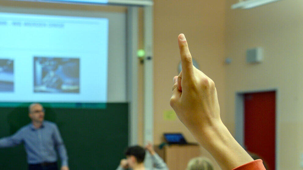 Blick von hinten in einen Klassenraum über die Köpfe der Schüler. Im Hintergrund steht ein männliche Person vor einer Tafel. Im Bildvordergrund ist der zum Melden gehobene Arm eines Schüler zu sehen.