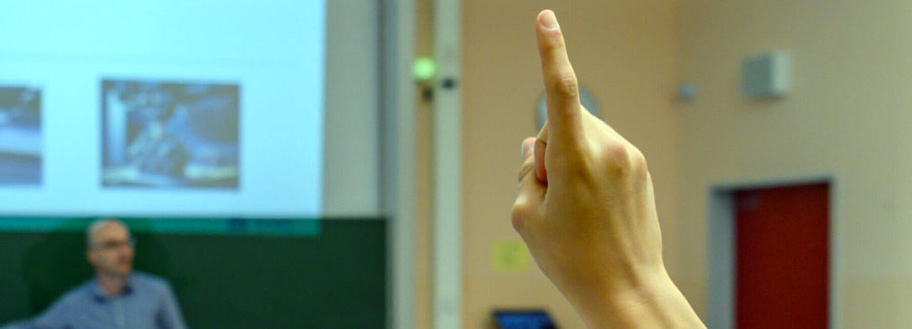 Blick von hinten in einen Klassenraum über die Köpfe der Schüler. Im Hintergrund steht ein männliche Person vor einer Tafel. Im Bildvordergrund ist der zum Melden gehobene Arm eines Schüler zu sehen.