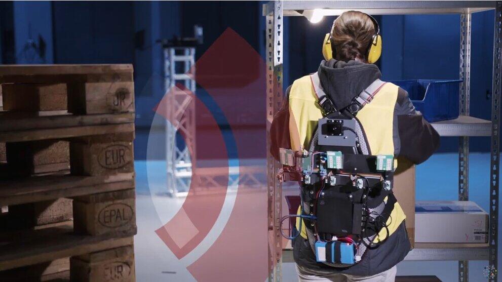 Person steht mit dem Rücken zur Kamera, trägt Weste mit technischer Ausstattung, Funkwellen gehen zu einer Palette