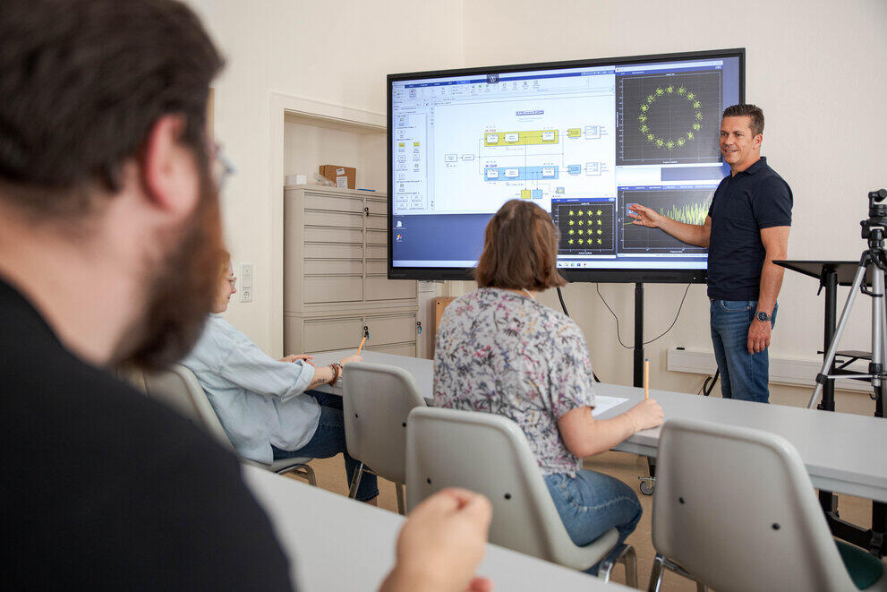 A lecturer is standing at a digital whiteboard, explaining something about modern transmission methods to a group of students seated in front of him.
