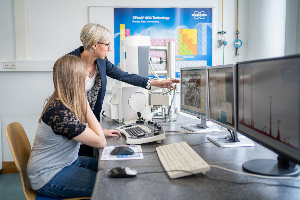 Eine Studentin und eine Dozentin stehen vor einem Computer und zeigen auf den Bildschirm.