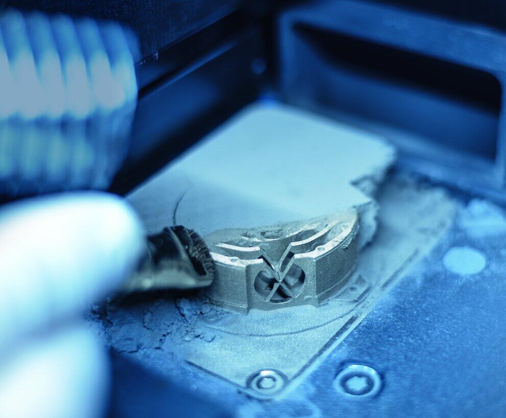 Ein aus Metall 3D-gedrucktes, sehr kleine Bausteil wird mit einem Pinsel vom überschüssen Metallpulver befreit.