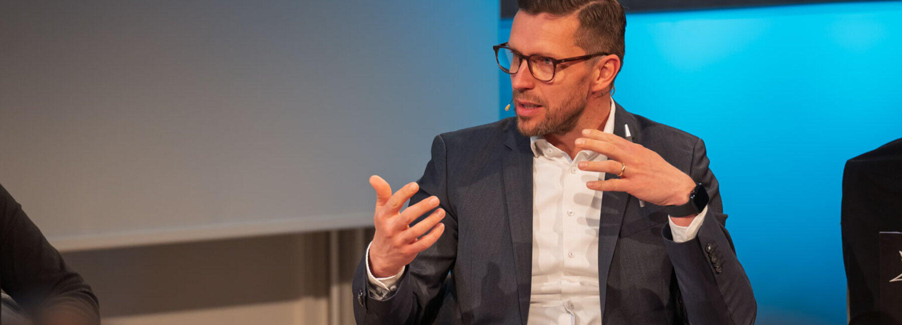 Professor Alexander Knauer is sitting on a panel and gesticulating in a relaxed manner.