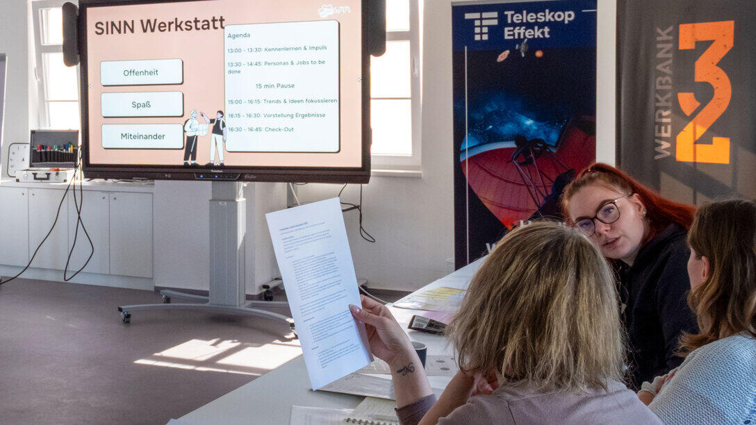 : Drei Personen im Gespräch. Im Hintergrund ist ein großer Screen zu sehen, auf dem „SINN Werkstatt“ zu lesen ist.