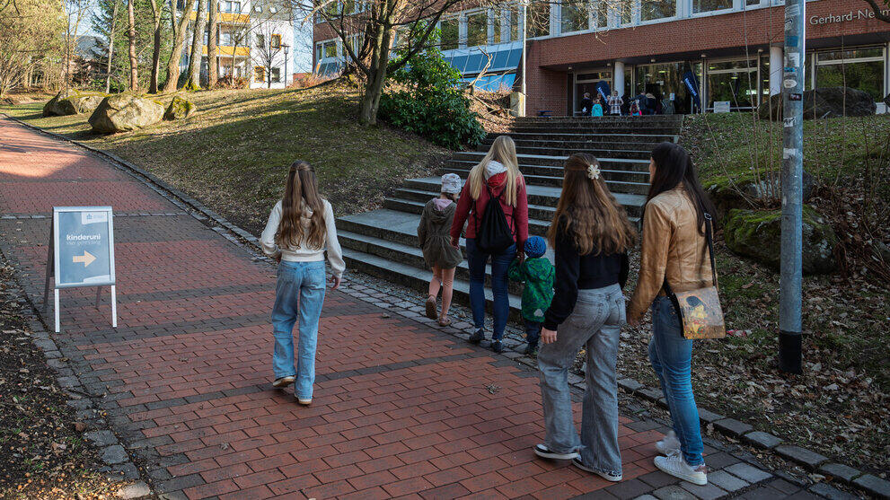 Kinder und Erwachsene auf dem Weg zum Hörsaalgebäude. Auf dem Weg steht ein Kinderuni-Aufsteller.