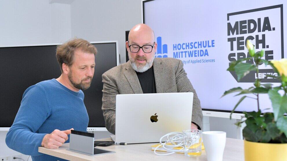 Two men are standing by a table, looking at a laptop screen.