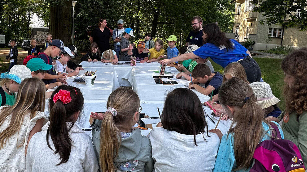 iele Kinder sitzen an Tischen unter einem Baum und arbeiten. Ringsum stehen Erwachsene.