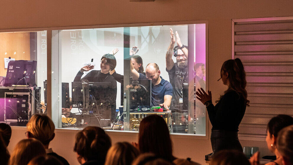 A view through a glass pane into the brightly lit control room, from which four people are waving cheerfully. In the foreground, the heads of the audience can be seen from behind.