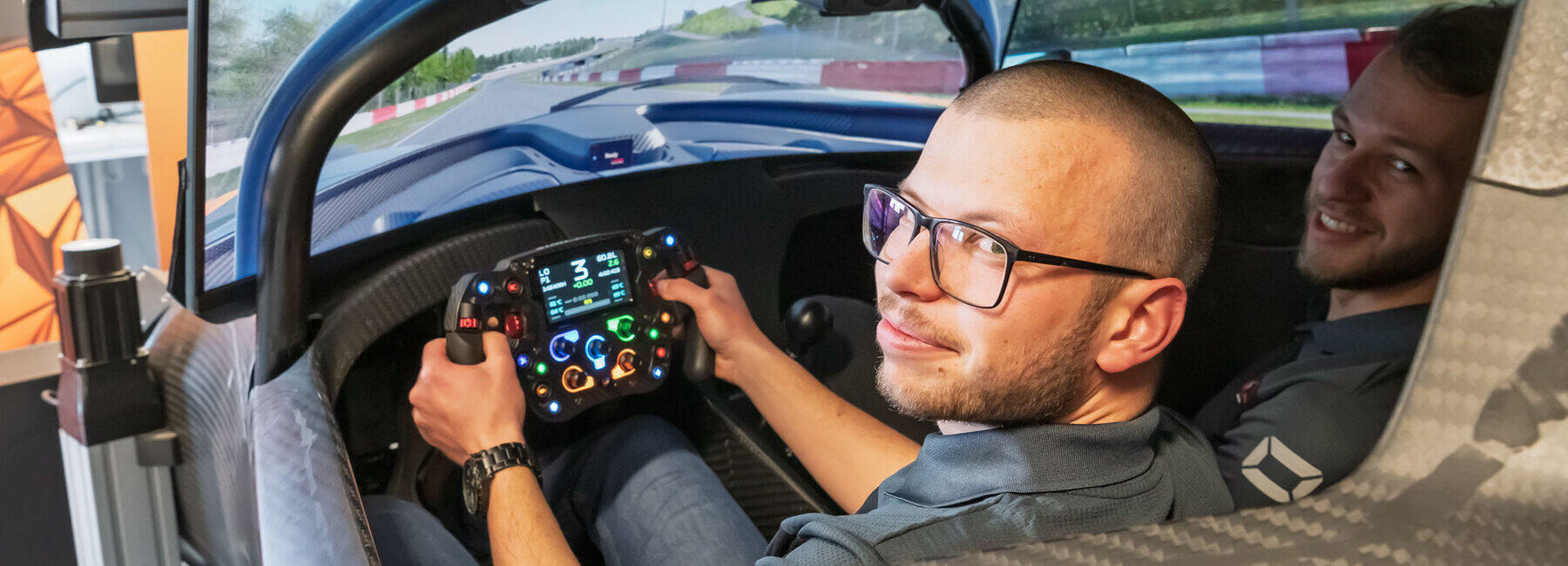 Zwei junge Männer im Cockpit eines Rennwagens, das Teil einer Simulationsplattform ist, in der das Umfeld Rennstrecke auf großen Monitoren dargestellt ist. Der linke Mann fasst mit beiden Händen das Lenkrad. Beide schauen nach hinten in Richtung Kamera.