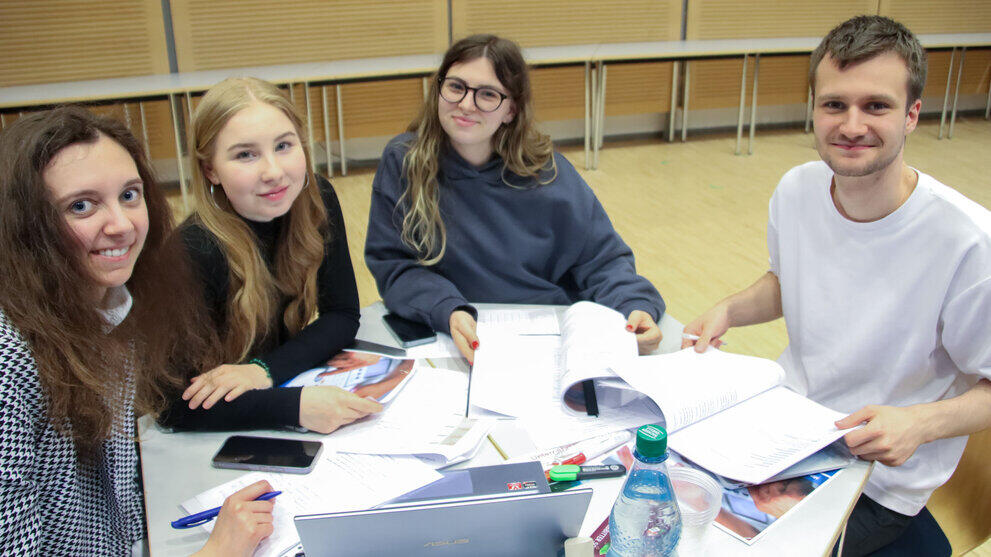 Three young women and one young man are sitting at a table with laptops and lots of papers, smiling at the camera.