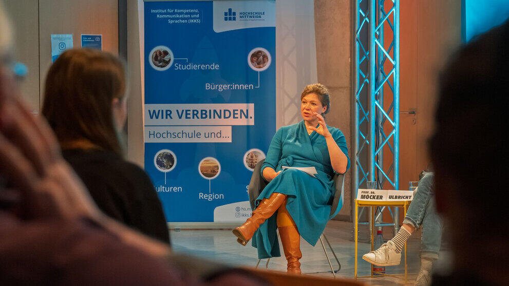 Weibliche Person sitzend zum Publikum sprechend. Köpfe von Zuschauenden unscharf von hinten zu sehen. Im Hintergrund ein blaues Rollup unter anderem mit dem Text "Wir verbinden Hochschule und ... ".