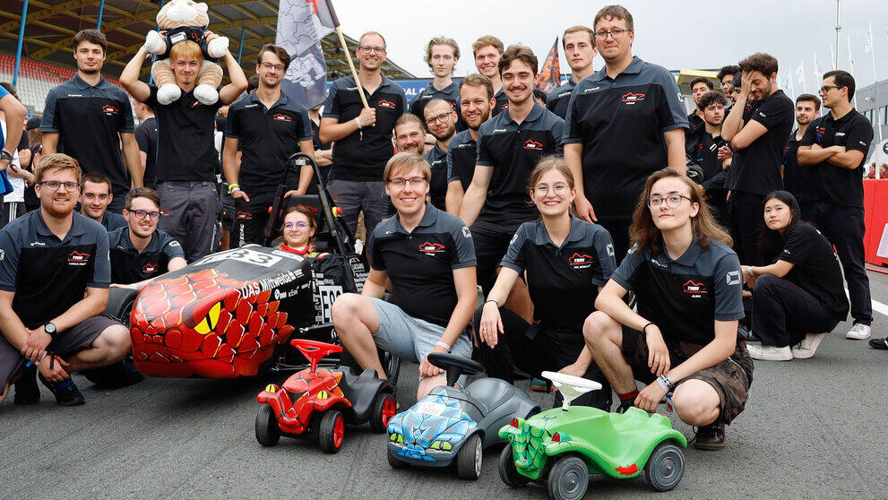 Team aus 20 jungen Menschen, das Rennauto und drei Bobbycars.