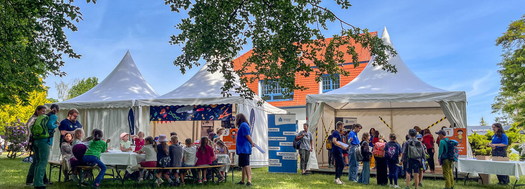 Drei Pavillions auf einer Wiese. Viele Kinder sitzen und stehen davor und darin.