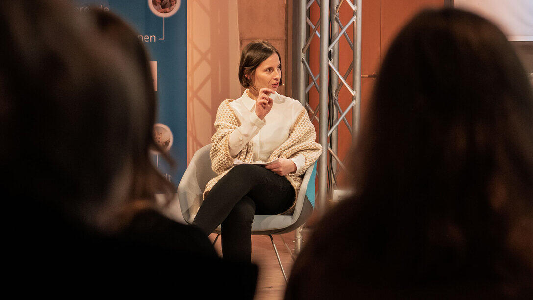 A view through the audience towards the presenter. She is gesturing and addressing the panellists, who are not visible in the shot.