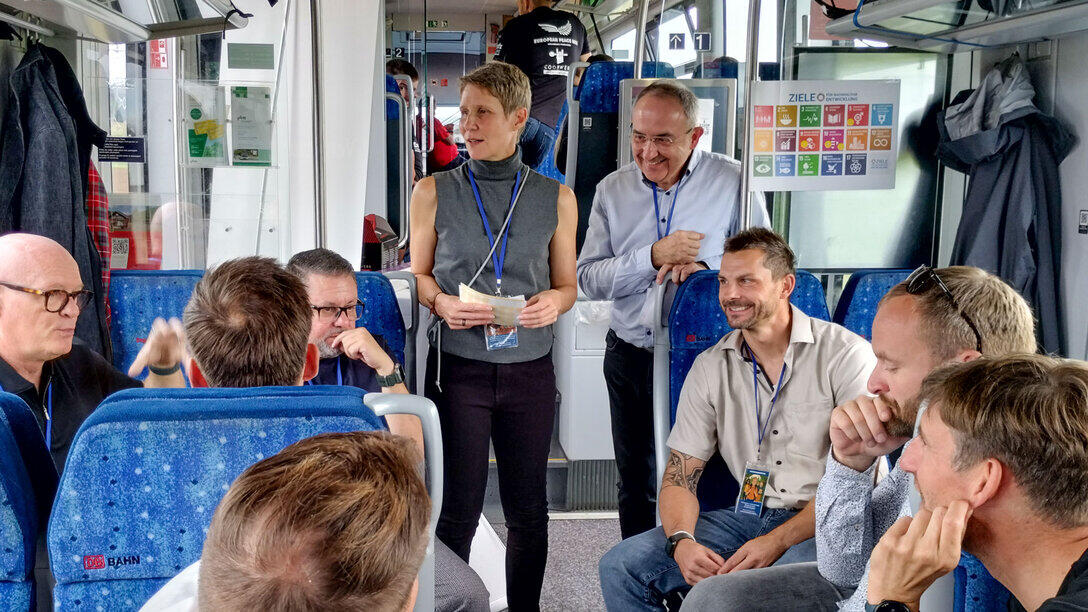 A group of people are sitting on a regional train, listening to two speakers standing in the aisle