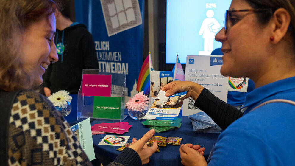 A woman hands a flyer with the heading ‘Diversity Management’ to another woman.