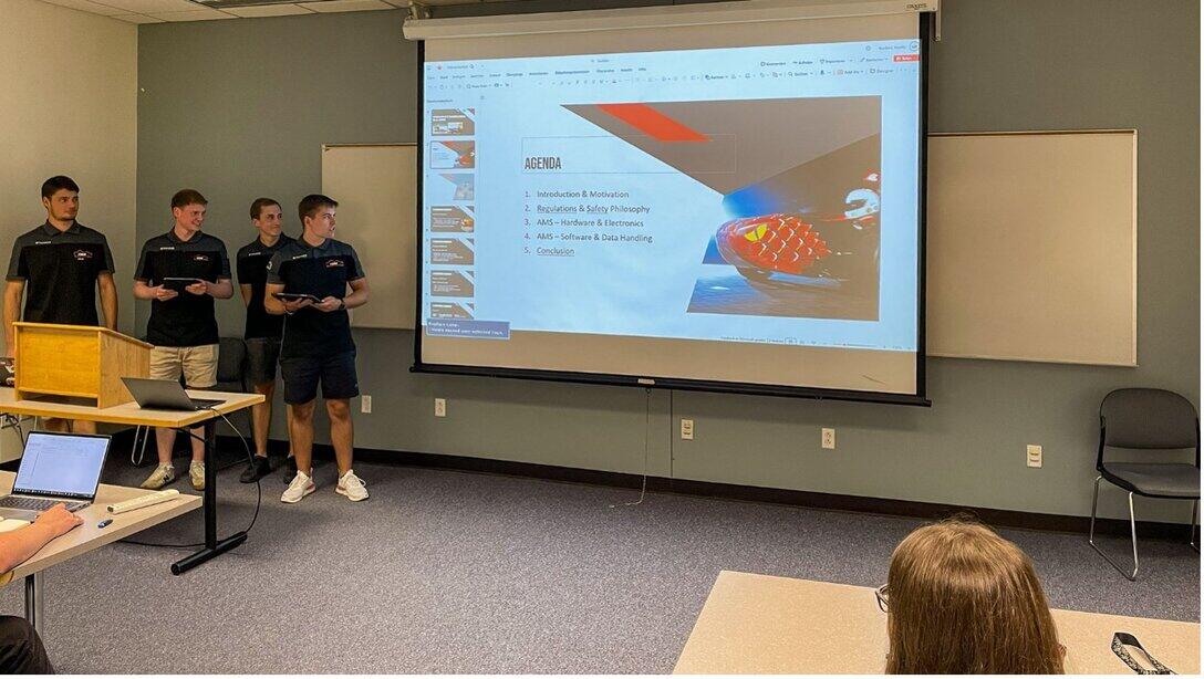 Four men are standing next to a screen displaying a PowerPoint presentation. The slide bears the heading ‘Agenda’ and shows the front end of a racing car.