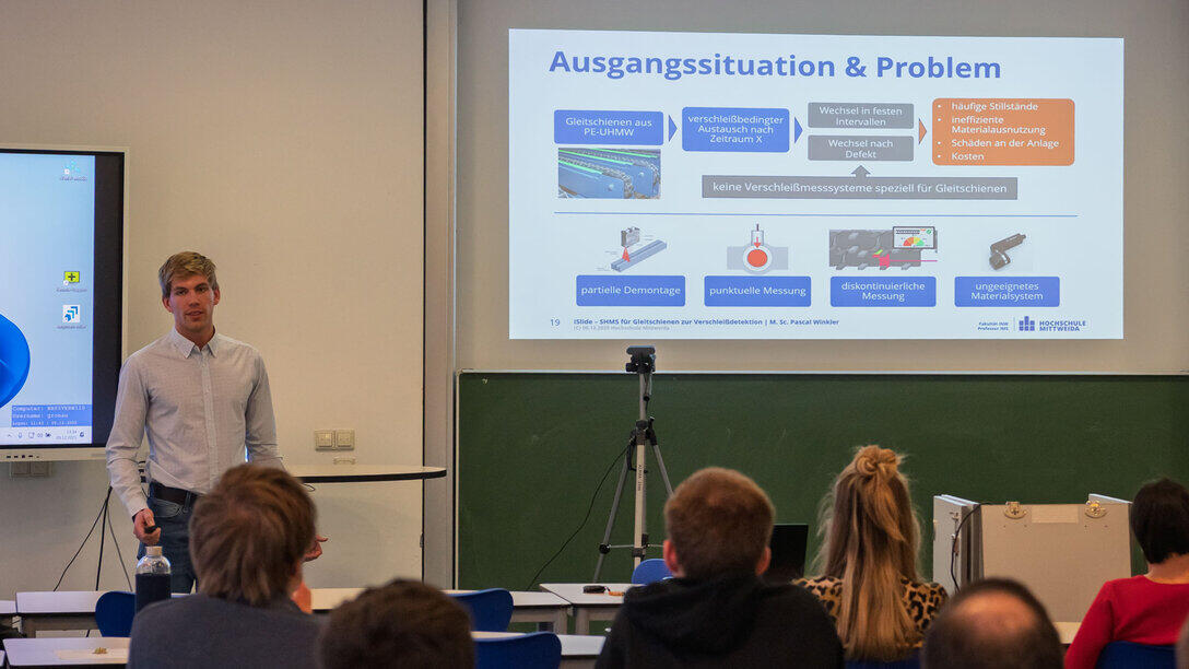 A man standing in front of a projected PowerPoint presentation. In the foreground, the backs of the audience’s heads.