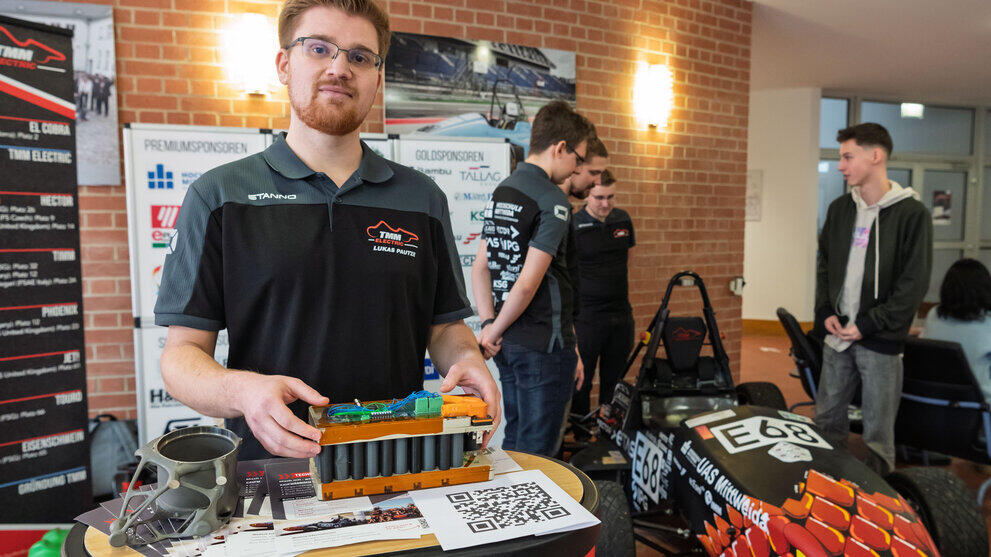 A young man wearing a ‘TMM’ polo shirt and carrying a battery pack. In the background, a group of men are standing around an (electric) racing car.
