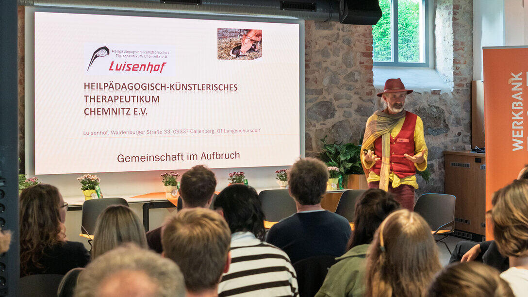 Eine männliche Person mit Hut spricht zum Publikum. Auf dem Screen links neben ihr ist unter anderem zu lesen „Luisenhof. Gemeinschaft im Aufbruch“.