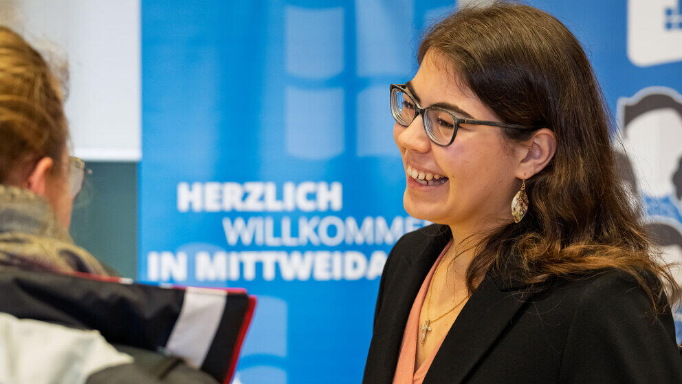A young woman laughing heartily whilst chatting. In the background, a roll-up banner reads ‘A warm welcome to Mittweida’.