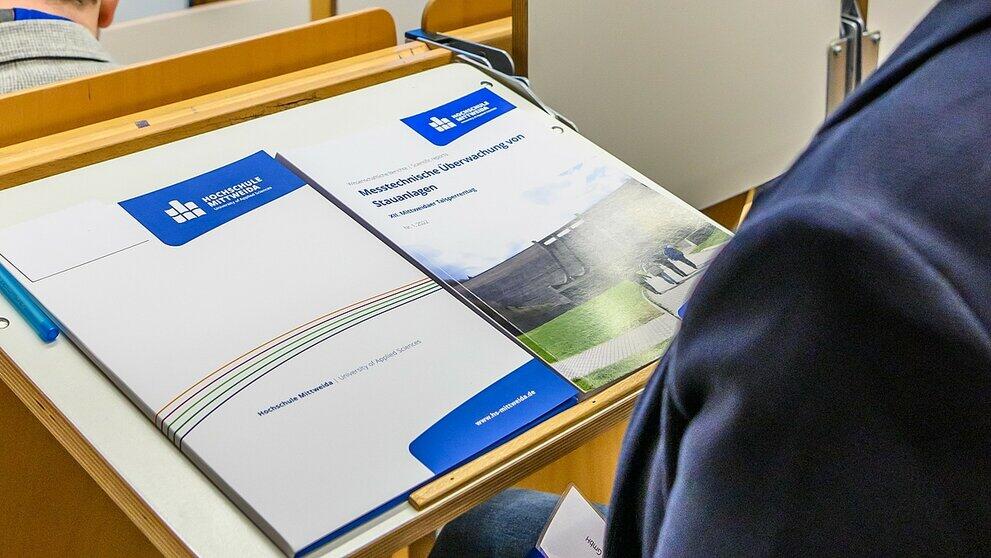 Blick auf einen Tagungsband und eine Mappe auf einem Klapptisch im Hörsaal