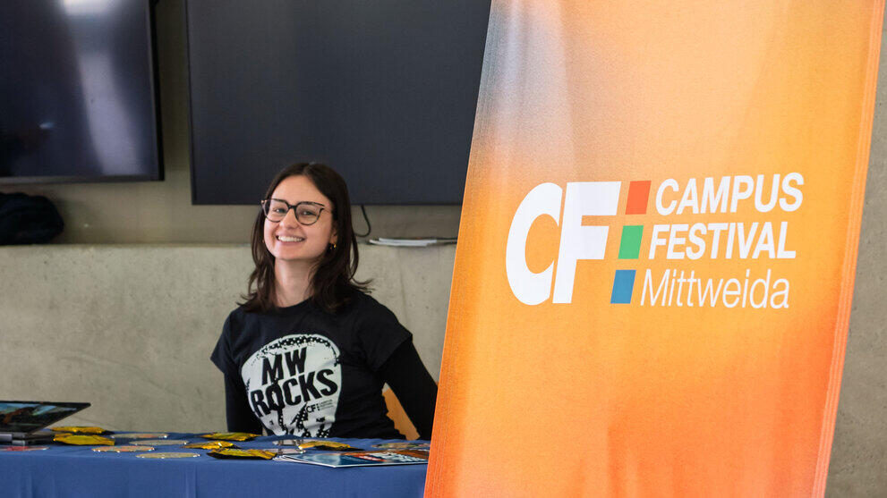 Eine junge weibliche Person mit "MW rocks"-Polo sitzt fröhlich lächeld an einem Tisch. Neben ihr im Bildvordergrund steh ein "Campusfestival Mittweida"-Rollup