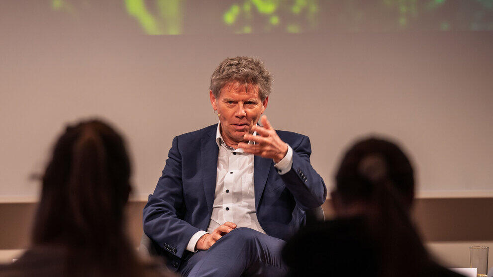Podiumsteilnehmer sitzend und ernst gestikulierend. Im Vordergrund sich unscharf Köpfe von Zuschauenden zu sehen.