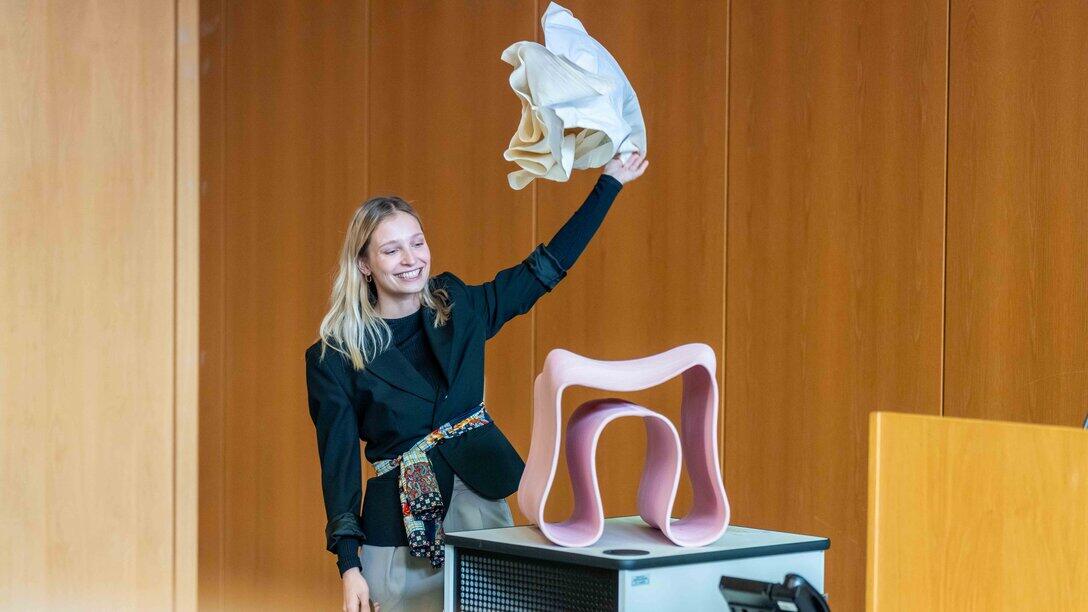 eine weibliche Person hält ein Tuch in Höhe und steht hinter einem Design-Stuhl