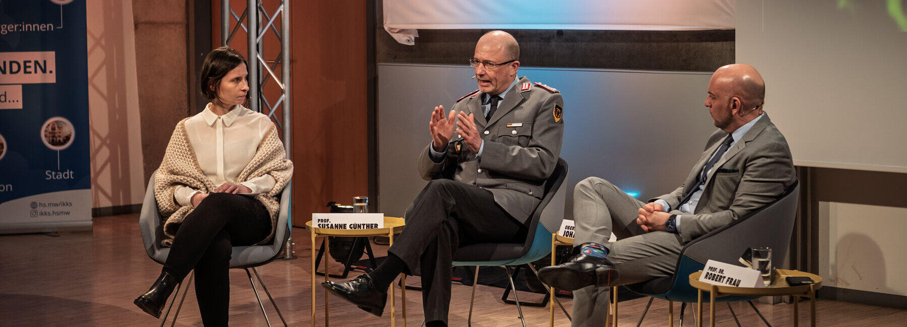Eine weibliche und zwei männliche Personen in Sesseln im Halbkreis. Die mittlere Person trägt eine Bundeswehruniform.