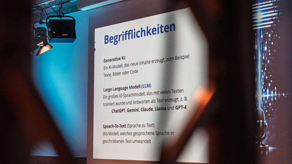 Blick auf eine projizierte Folie mit der Überschrift „Begrifflichkieten“ und den erläuternden Anstrichen: „Generative KI: Ein KI-Modell, das neue Inhalte erzeugt, zum Beispiel Texte, Bilder oder Code; Large Language Modell (LLM): Ein großes KI-Sprachmodell, das mit vielen Texten trainiert wurde und Antworten als Text erzeugt, z. B. ChatGPT, Gemini, Claude, Llama und GPT-4; Speach-To-Text (Sprache zu Text): (KI)-Modell, welches gesprochene Sprache in geschriebenen Text umwandelt.“