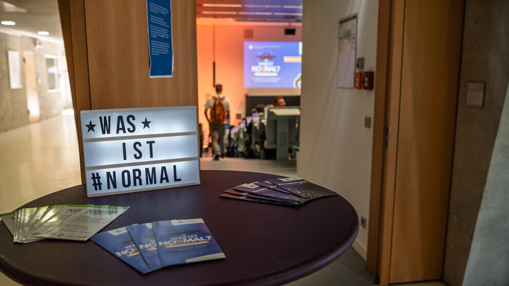 Auf einem runden Stehtisch vor einer geöffneten Hörsaaltür sind vor einem Leuchtschild mit dem Text „Was ist normal“ Flyer ausgebreitet.
