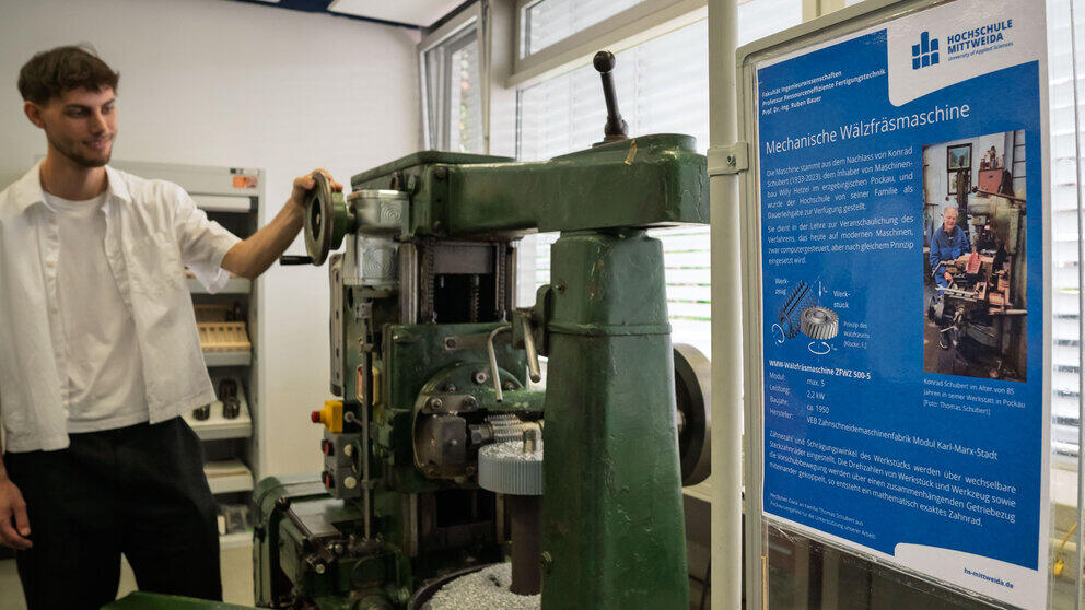 In the foreground on the right of the picture is a poster bearing the HSMW logo, which, against a blue background, explains the function and history of the machine in the centre of the picture. At the left-hand edge of the picture stands a young man who has rested his left hand on one of the machine’s control wheels and is looking at it.