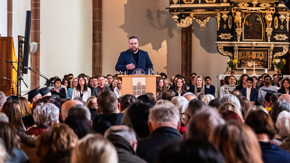 A view over the heads of the congregation seated in the pews of the nave, towards a man at the lectern in the background.
