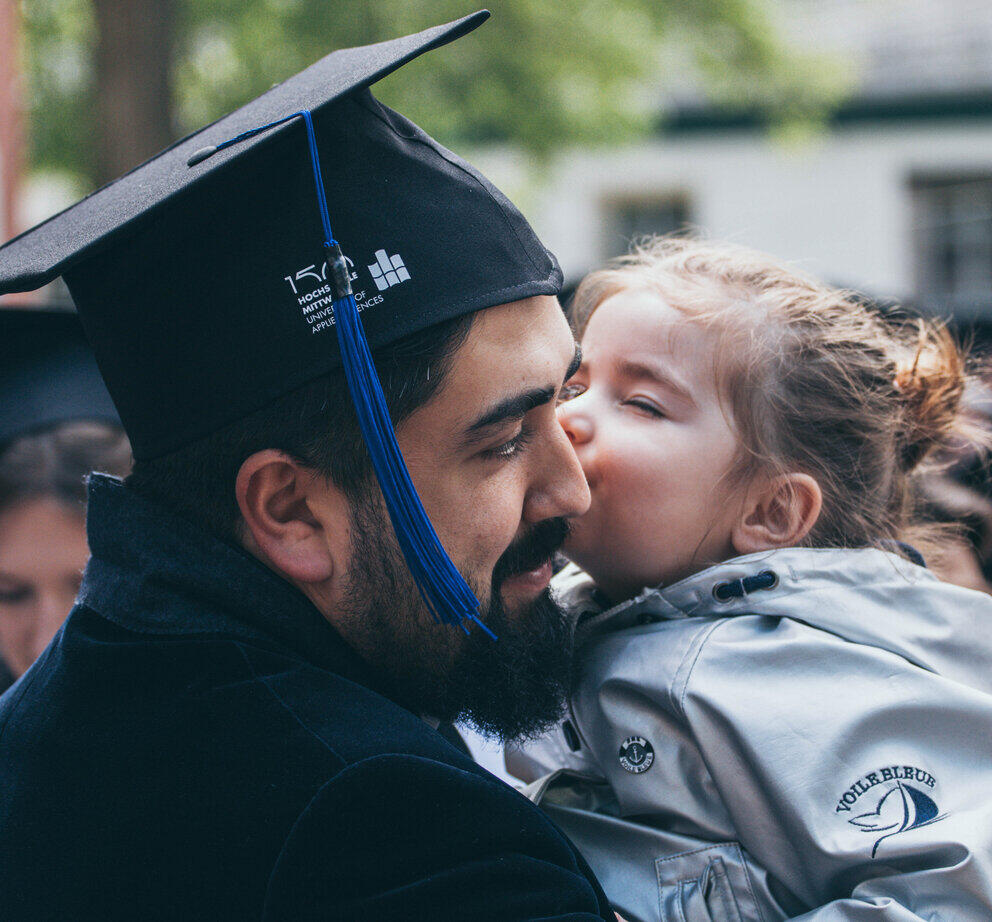 Ein Mann mit Graduiertenhut wird von seiner Tochter im Kindesalter geküsst. Er hält sie auf dem Arm.
