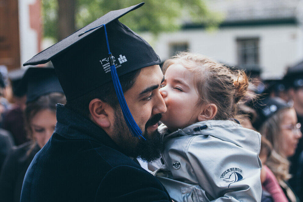 A man wearing a graduation cap is being kissed by his young daughter. He is holding her in his arms.