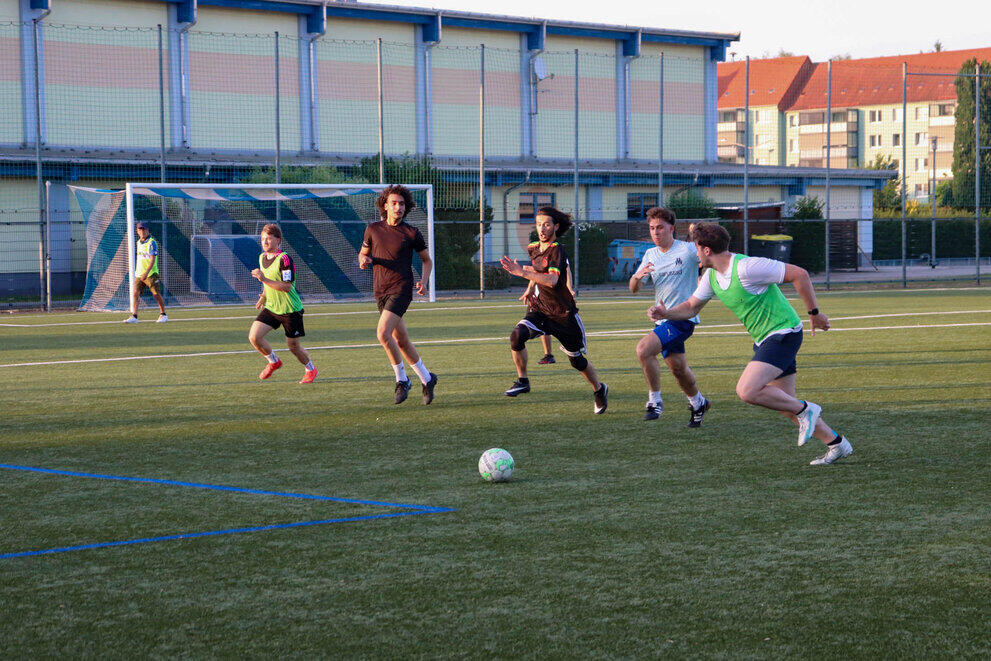 Sechs Personen spielen auf einem Kunstrasenplatz in zwei Teams Fußball.