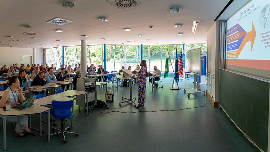 A wide shot of the event room. On the left-hand side of the frame, the participants are seated at tables. Facing them, a speaker stands at the lectern. At the right-hand edge of the frame, a projected slide with the heading ‘Introduction’ can be seen, with illegible text embedded within graphic elements below it.
