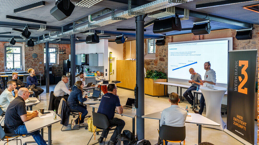 A room with exposed brickwork, metal columns and surface-mounted wiring. Two men stand in front of a large screen. Opposite them, several people are seated, mostly at tables, listening.