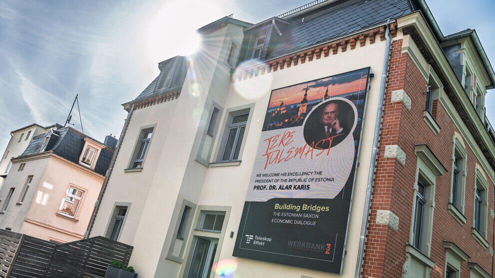 Eine Hausfassade mit einem wandfüllenden LED-Display: Es zeigt das Foto von Alar Karis und unter anderem den Text „Building Bridges“. Im gegen die Sonne gemachten Foto spiegeln sich die Linsen des Objektivs.
