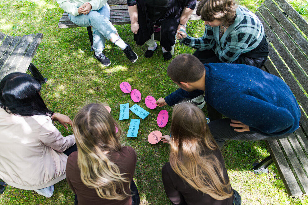 Mehrere Studierende sitzen während einer Gruppenarbeit auf Bänken auf einer Wiese im Kreis und legen beschriftete Zettel in die Mitte.