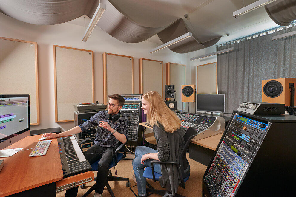 Zwei Studierende sitzen in einem Tonstudio mit viel Technik und Mischpulten und schauen auf einen Computerbildschirm.