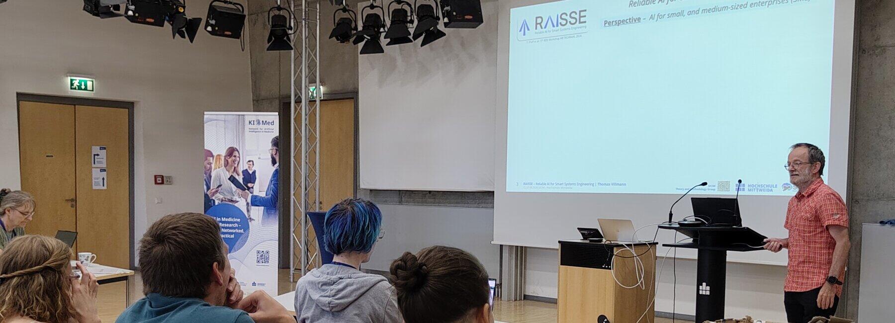 Person steht vor Präsentationswand, Zuhörende sitzen an Tischen
