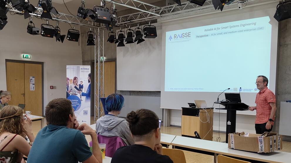Person steht vor Präsentationswand, Zuhörende sitzen an Tischen