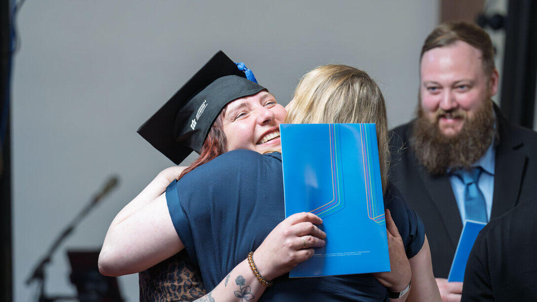 Eine Absolventin mit Graduiertenhut wird von ihrer Professorin umarmt.