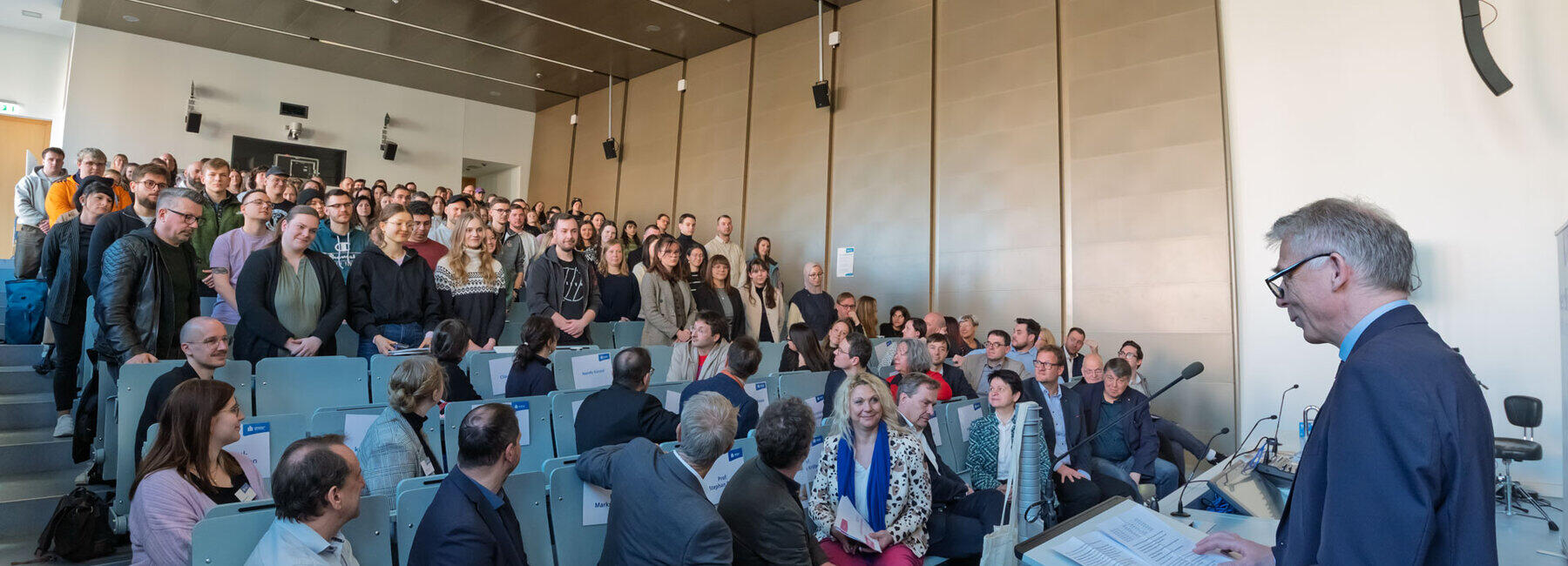Young people standing in the rising rows of seats in a lecture theatre. Older people are seated in the lower rows and are turning to look at them.